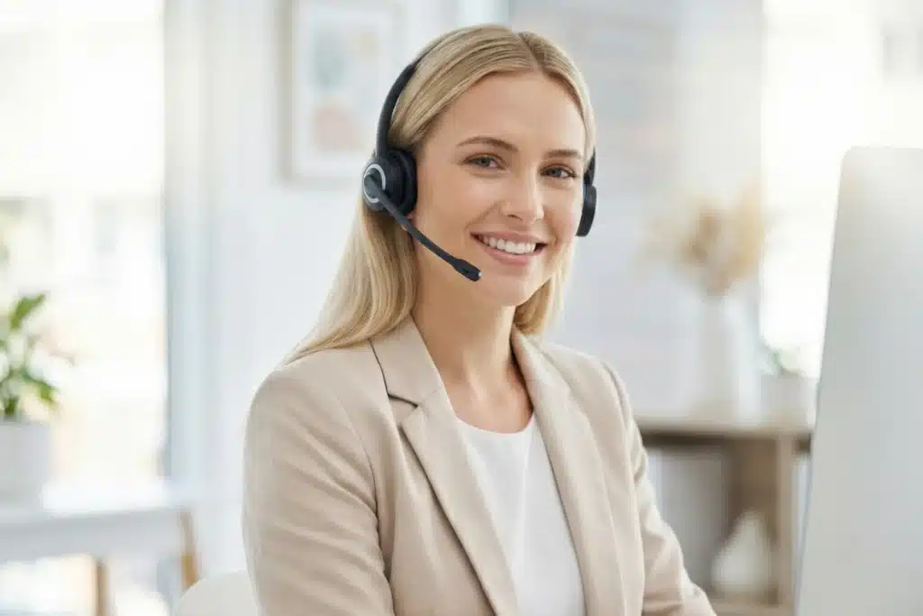 Conseillère du service client portant un casque micro, souriante et installée à son poste dans un bureau lumineux.