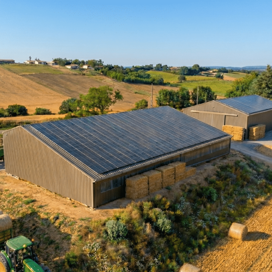 Hangars agricoles équipés de toitures photovoltaïques, situés au cœur d’une exploitation rurale avec tracteurs, bottes de foin et champs récoltés.
