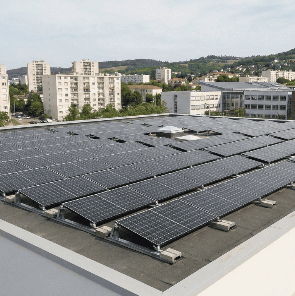 Installation photovoltaïque en toiture sur un bâtiment public, composée de plusieurs rangées de panneaux solaires plats, avec une zone urbaine et des immeubles en arrière-plan.